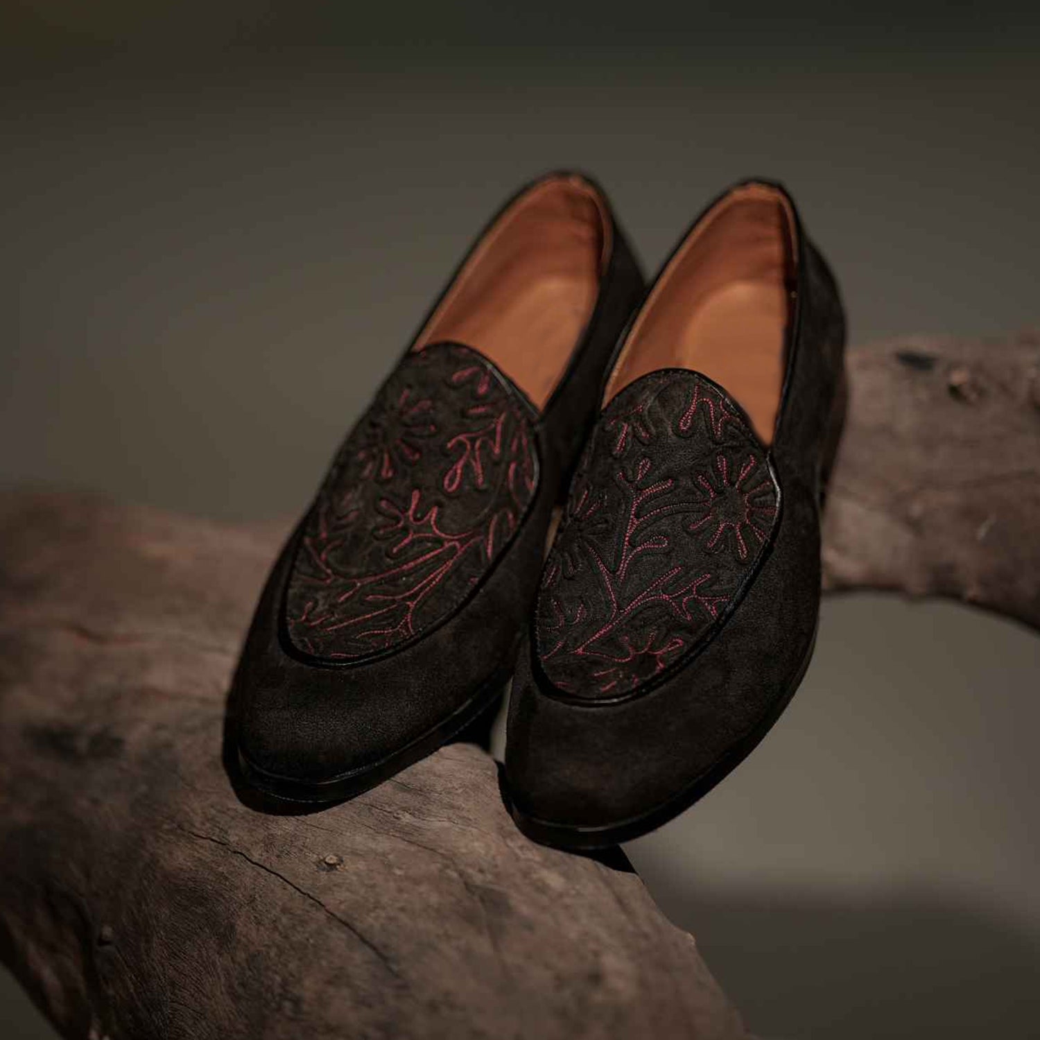 Pair of black leather loafers with embroidered floral pattern placed on a rustic wooden branch.