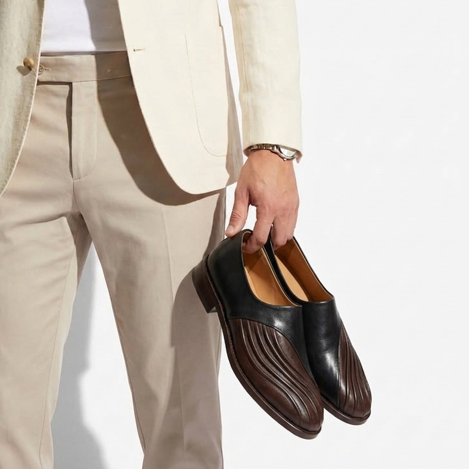 Pair of elegant two-tone leather slip-on shoes with black uppers and brown pleated toe detail, displayed on a pedestal against a neutral background.