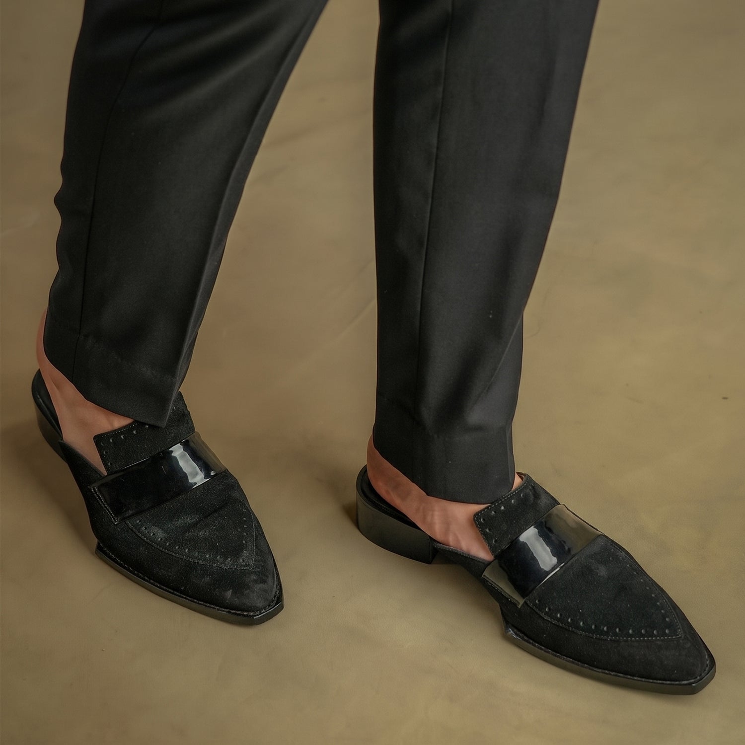 Pair of black suede slip-on mule loafers for men with pointed toe design, glossy strap detail, open-back style, and low block heel displayed on a neutral background.