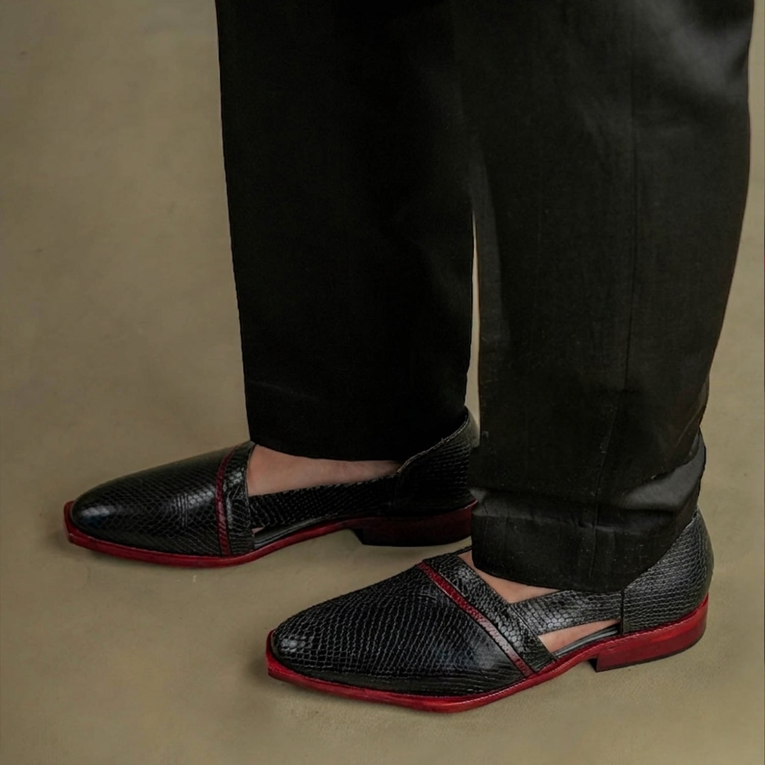 Pair of black textured slip-on mule loafers for men with reptile pattern finish, maroon edge detailing, pointed square toe, and open-back design placed on a wooden surface.