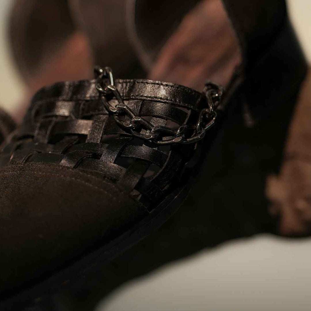 Men wearing black loafers with a woven leather upper and textured matte front, featuring a stylish metal chain detail across the vamp. The slip-on shoes have a sleek, modern silhouette paired with black trousers, set against a neutral background, highlighting their premium design and sophisticated finish.
