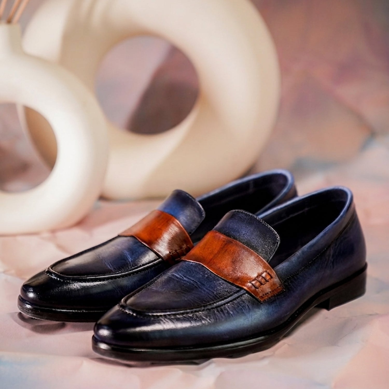 Close-up of navy blue leather penny loafers with a brown strap detail, worn with tailored navy trousers, showcasing a polished two-tone design and slip-on formal footwear style.
