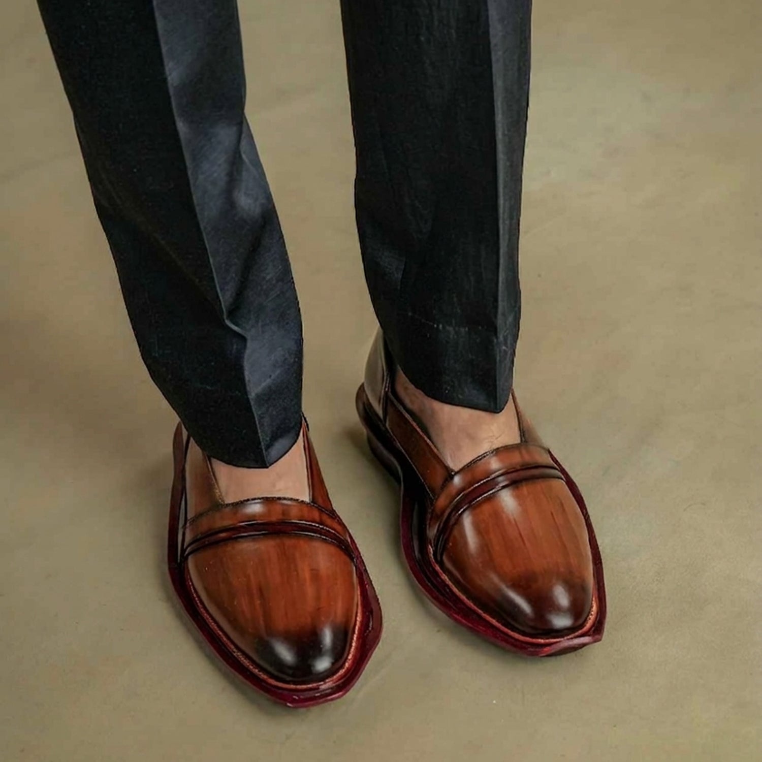 Pair of tan burnished slip-on mule loafers for men with almond toe design, strap detail across the vamp, maroon sole border, and open-back style displayed on a neutral background.
