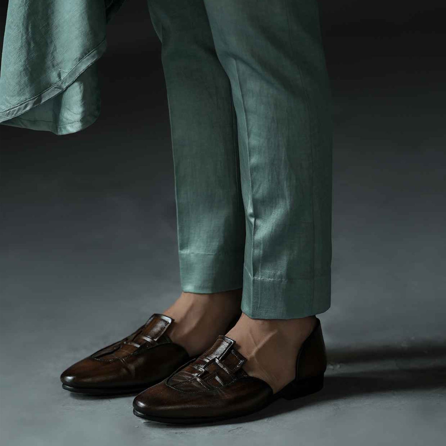 Person wearing brown leather loafers with buckle detail paired with cream trousers on a light floor.