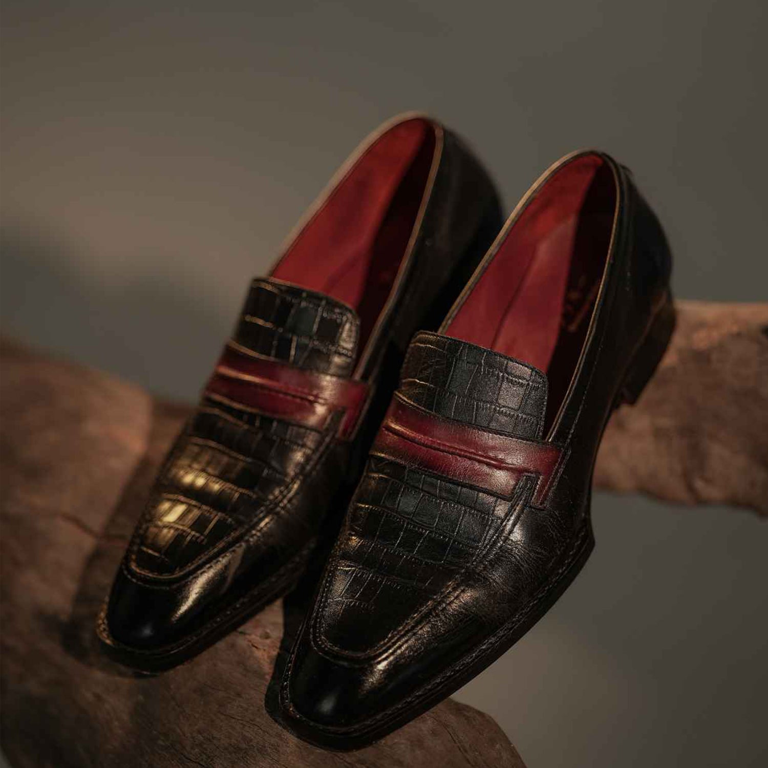 A pair of black men’s penny loafers placed on a wooden surface, featuring a crocodile-textured leather finish with a contrasting maroon strap across the front. The shoes have a polished appearance, a slightly elongated toe shape, and a vibrant red inner lining visible inside. Designed for formal and semi-formal wear, these slip-on loafers combine classic style with modern detailing.