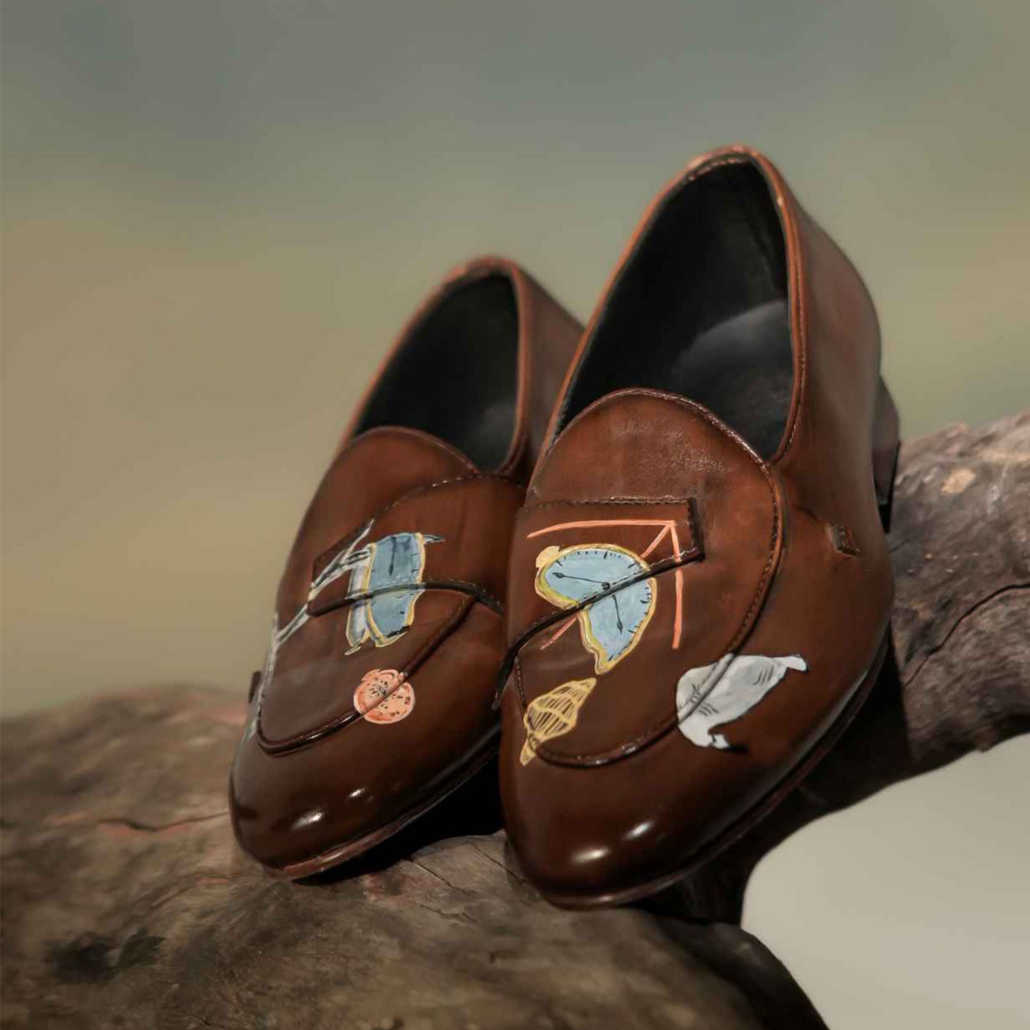 Pair of brown leather penny loafers for men with hand-painted abstract artwork, burnished finish, almond toe design, and slip-on style displayed on a rustic wooden surface.