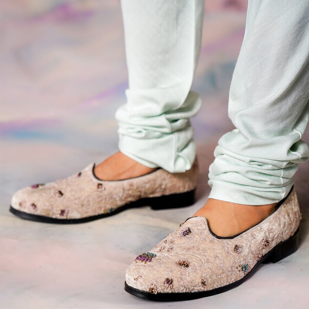 Men’s beige velvet embroidered loafers with intricate floral detailing, slip-on design, black sole, paired with light-colored trousers, perfect for ethnic and festive wear.