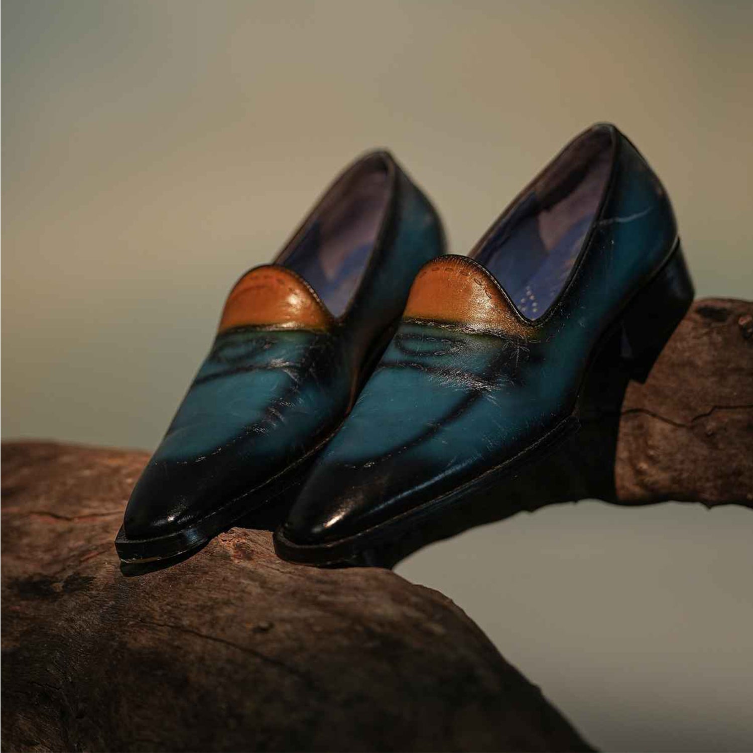 A pair of premium dual-tone leather slip-on shoes in teal and brown, featuring a polished finish and sleek elongated toe design. The shoes are crafted from high-quality leather with a smooth gradient effect, blending deep teal shades with warm brown accents near the opening. Positioned on a wooden surface, the shoes showcase a modern yet classic silhouette with a low heel and clean stitching. Ideal for formal and semi-formal wear, these stylish slip-ons offer a refined and sophisticated look for men.